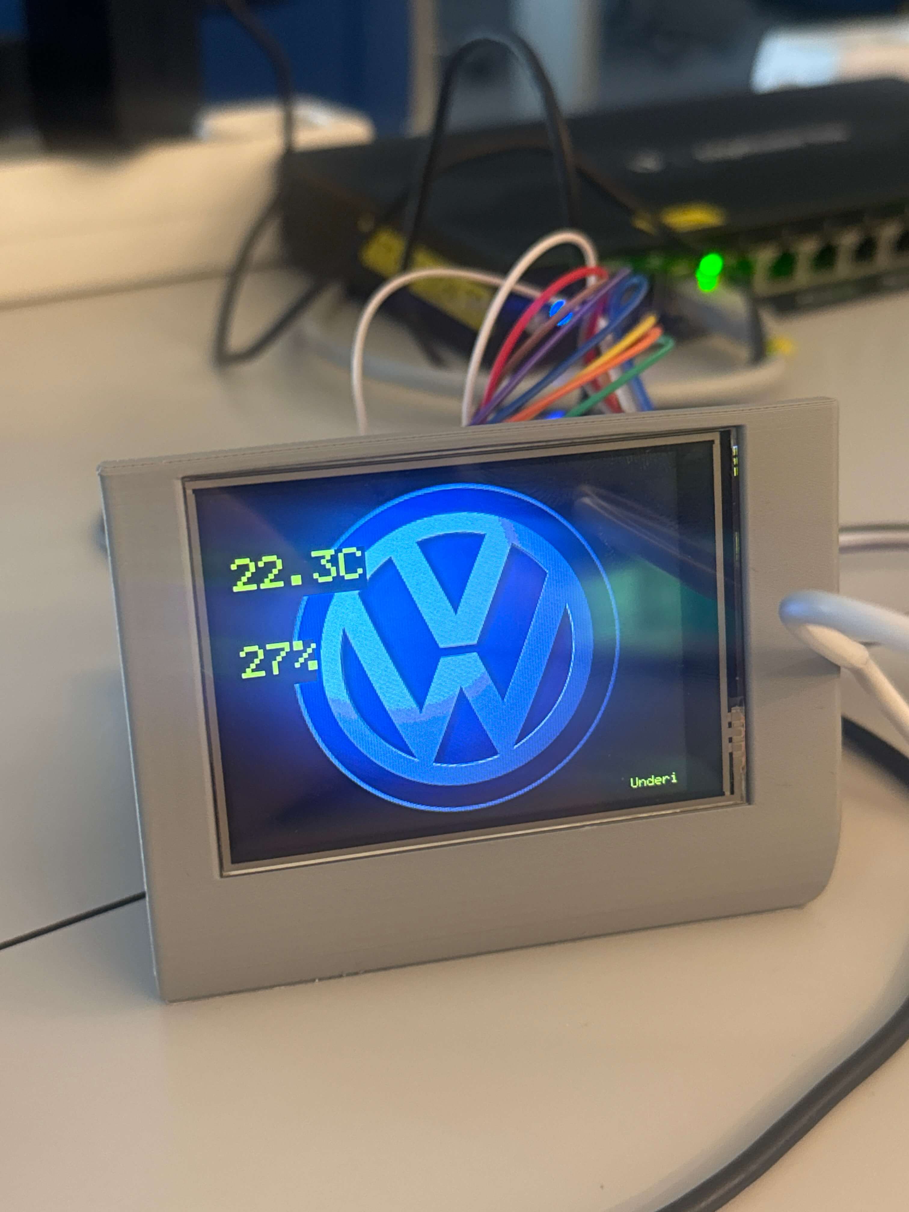 STM32 based automotive gauge display showing a large blue Volkswagen logo centered on the screen, two yellow green readings at top left reading 22.3C and 27%, and small green status text Underi at bottom right. The display is housed in a gray 3D printed pod resting on a desk with multicolored wires and a network switch blurred in the background. Technical prototype tone, focused and functional.